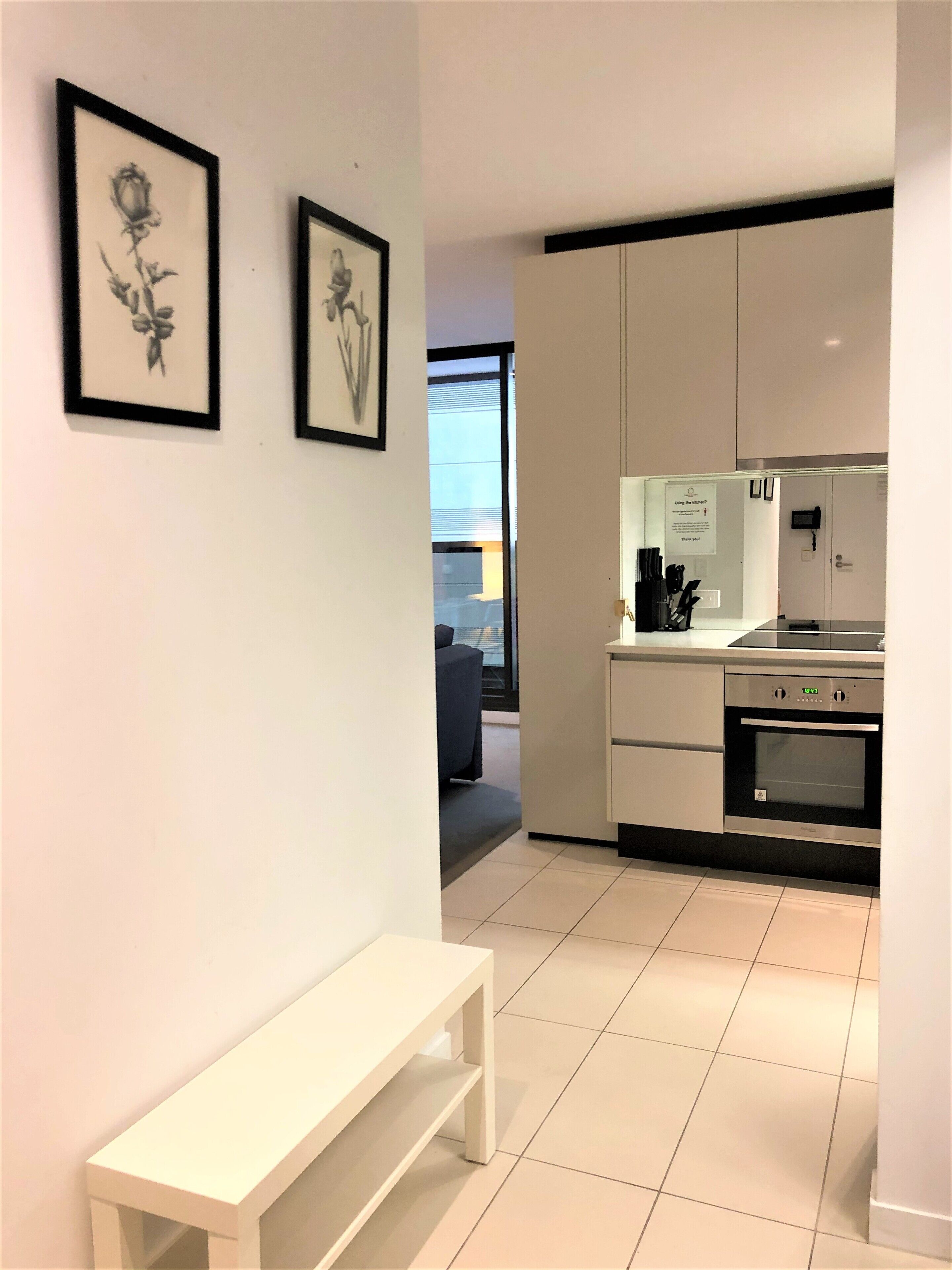 A modern kitchen with white cabinets, a built-in oven, and a digital clock. There is a bench in the foreground and framed pictures on the wall.