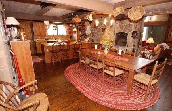 A rustic dining area with a wooden table and chairs, a stone fireplace, and a kitchen in the background.