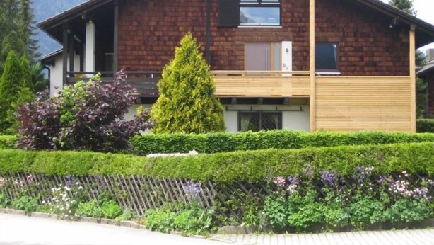 A two-story house with a wooden deck, a large window, and a well-maintained garden with various plants and a fence. - Maisonette apartment with mountain panorama for up to 5 people, 65 sqm, 1 bedroom (Bayerisch Gmain)