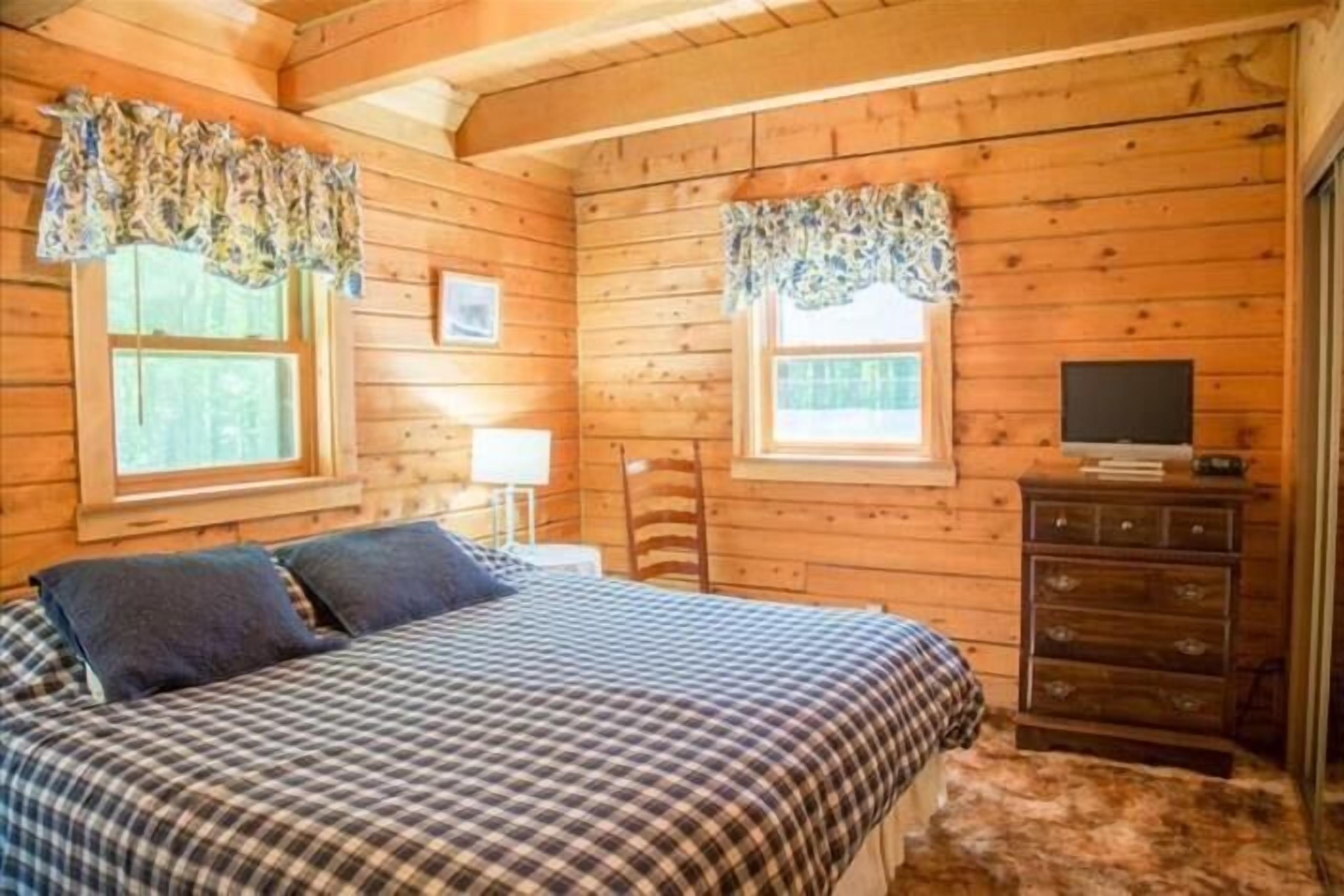 A wooden cabin bedroom with a bed, dresser, and two windows with curtains.