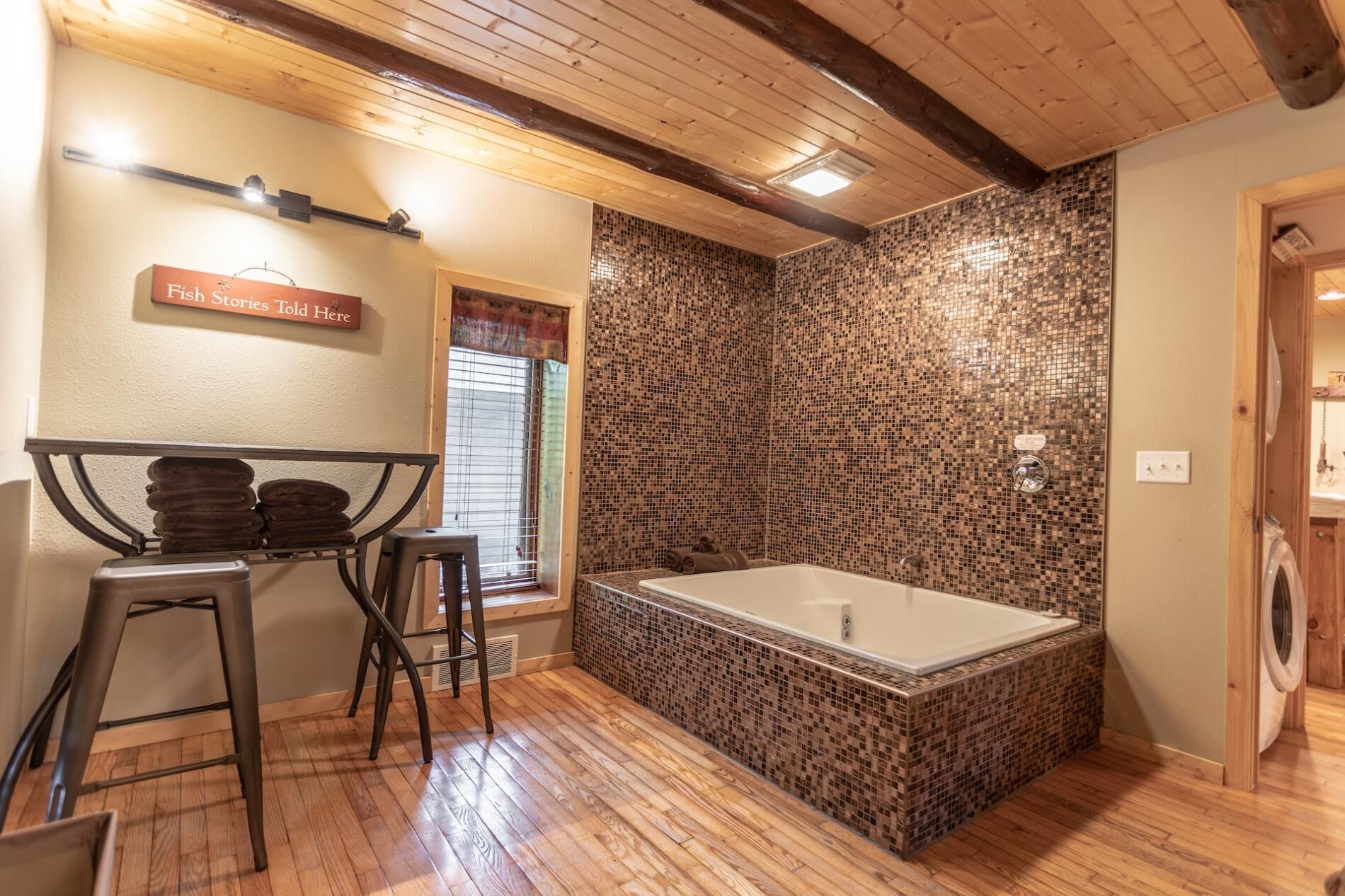 A bathroom with a mosaic-tiled wall, a wooden floor, a bathtub, and a sign that reads 'Fish Stories Told Here'.