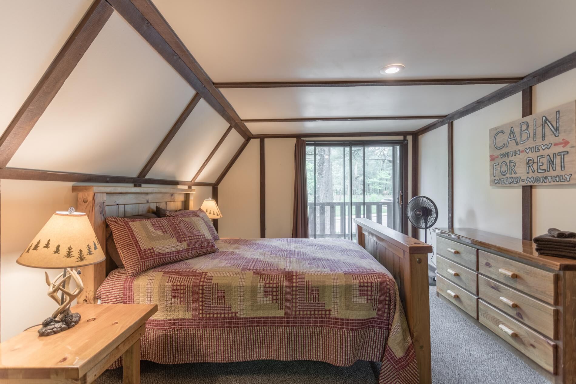 A bedroom with a sloped ceiling, a bed with a patterned quilt, a nightstand with a lamp, a dresser, and a sign that reads "CABIN WITH VIEW FOR RENT WEEKLY-MONTHLY."