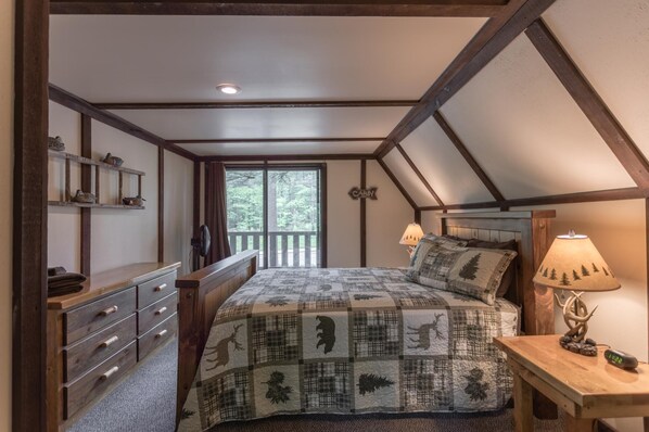 A bedroom with a sloped ceiling, a bed with a patterned quilt, a nightstand with a lamp, a dresser with drawers, and a window with a view of trees.