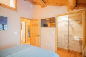 Quarto com cama, vigas de madeira no teto, guarda-roupa e banheiro com chuveiro e suporte para toalhas.