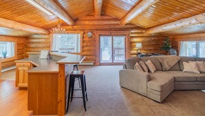 L’intérieur d’une cabane en rondins, avec un plafond en bois, un coin cuisine doté d’un bar, un canapé et une grande fenêtre.