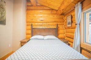 Ein Schlafzimmer in einer Blockhütte mit einem hölzernen Kopfteil, einer gemusterten Bettdecke, einem Nachttisch und einem Fenster mit Vorhängen.