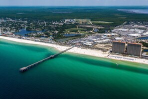 Vue aérienne d’une zone côtière avec une longue jetée s’étendant dans des eaux turquoise, une plage de sable et un grand complexe hôtelier composé de plusieurs bâtiments.