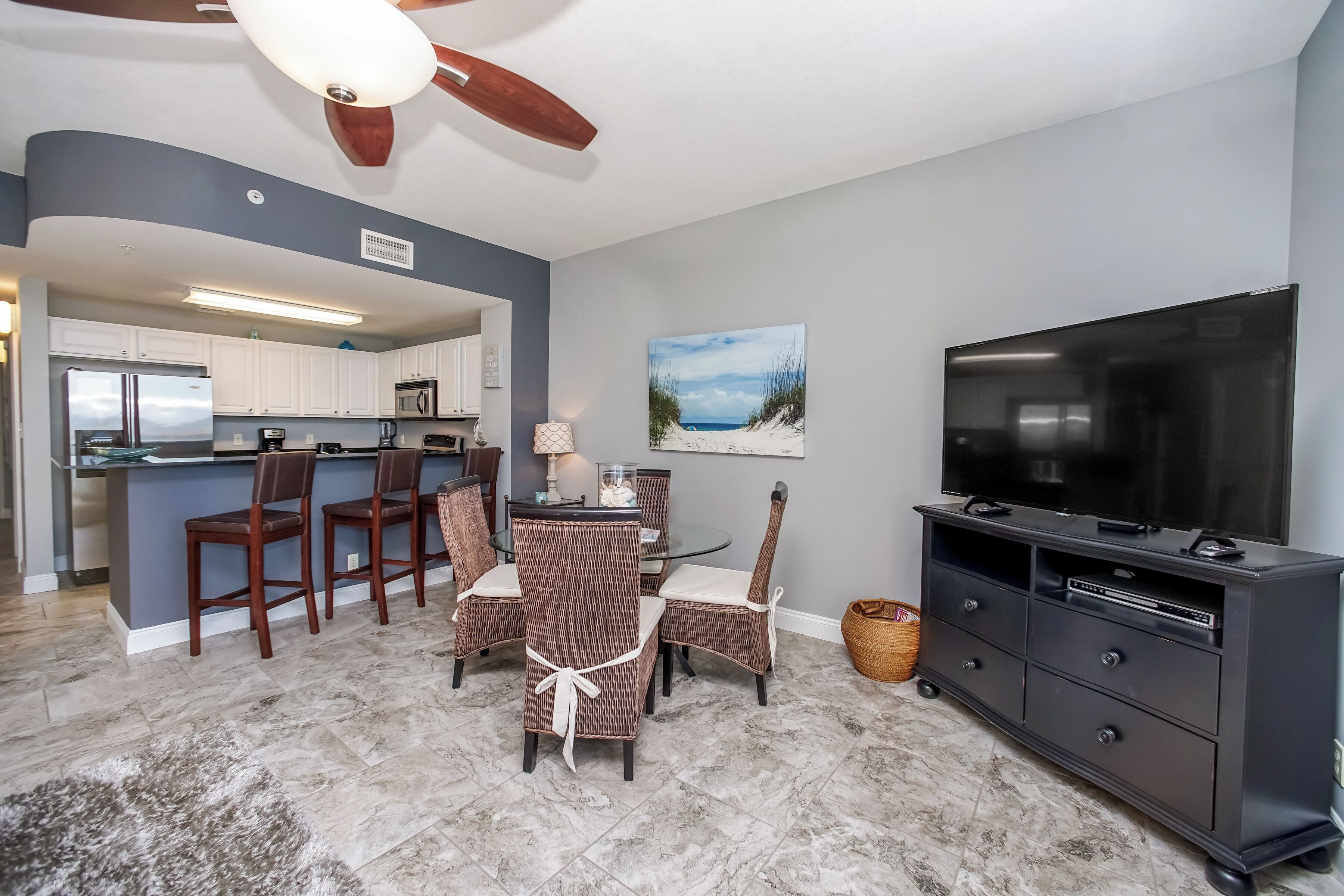 A modern kitchen with white cabinets, a dining area with wicker chairs, a flat-screen TV, and a ceiling fan.