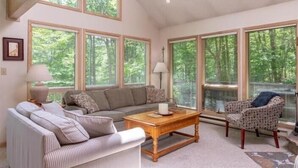 En hyggelig stue med sofa, lænestole og sofabord, store vinduer med persienner og et loft med synlige bjælker.