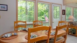 Et spiseområde med et træbord og stole, store vinduer med persienner, og et amerikansk flag kan ses gennem persiennerne.
