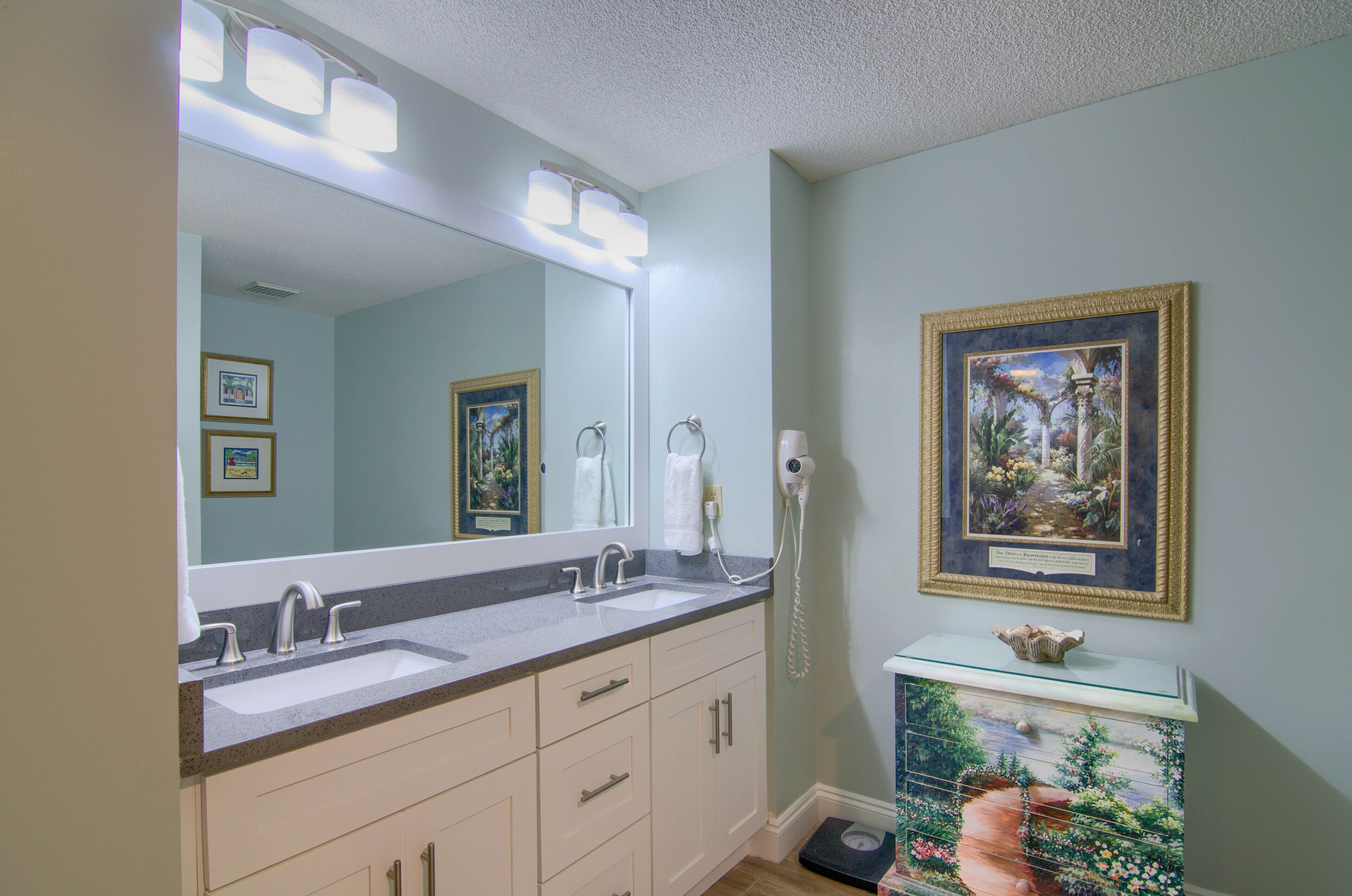 Une salle de bain avec un grand miroir, deux lavabos, une commode avec une balance et un tableau encadré au mur.