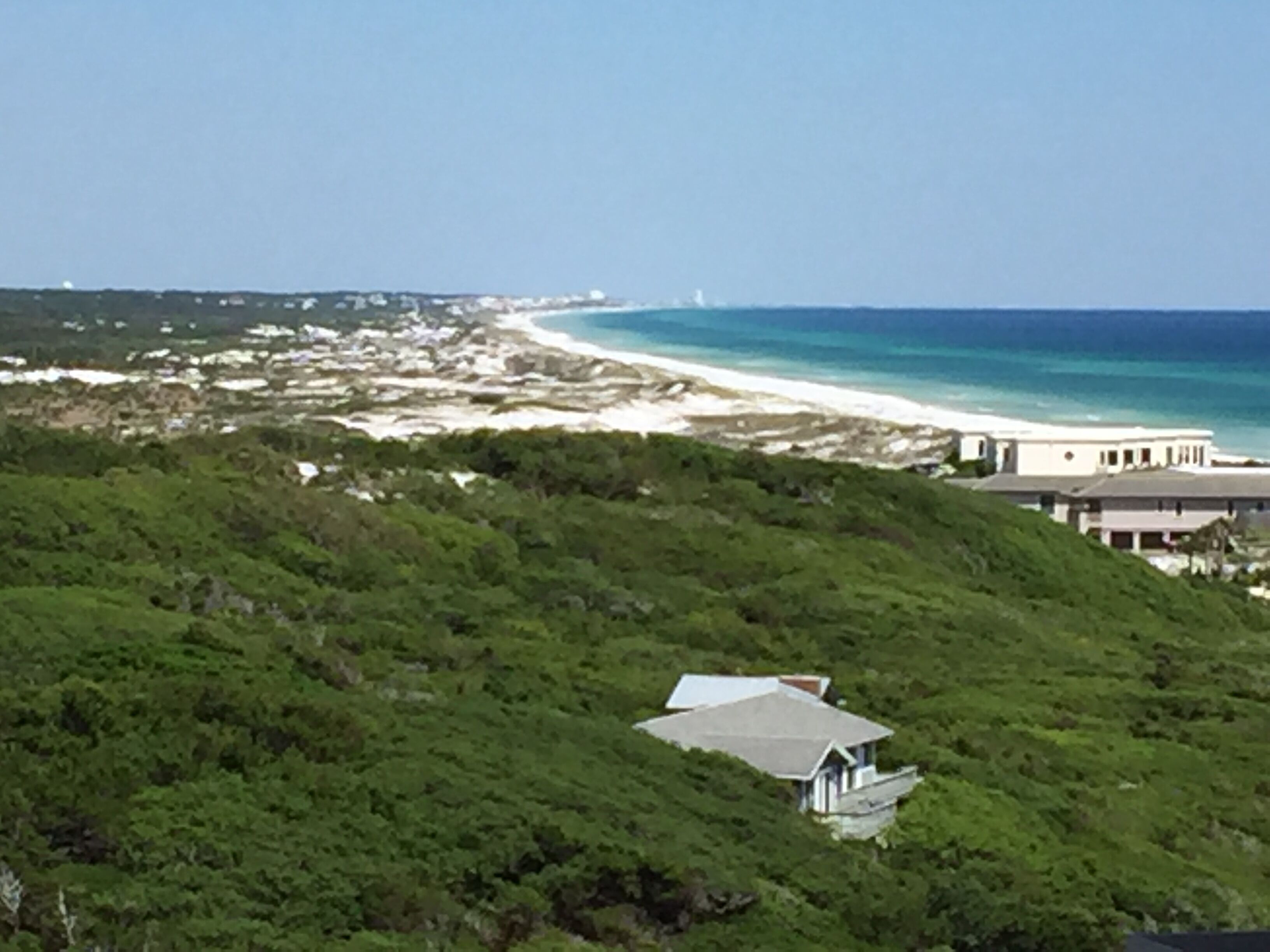 Vista costera con una playa, una casa en la colina y un faro a lo lejos.