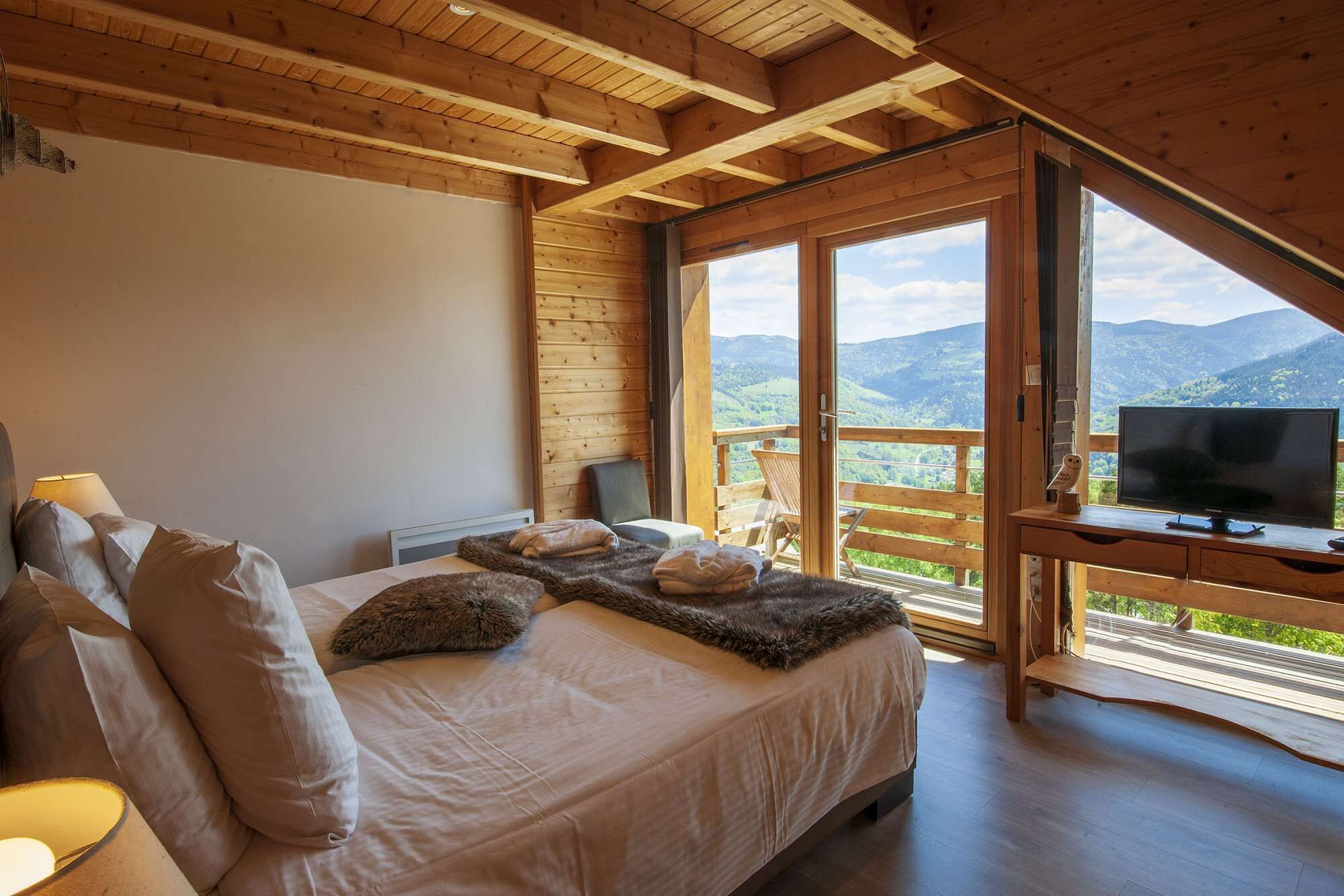 A bedroom with a bed, pillows, a TV, and a balcony with a mountain view.