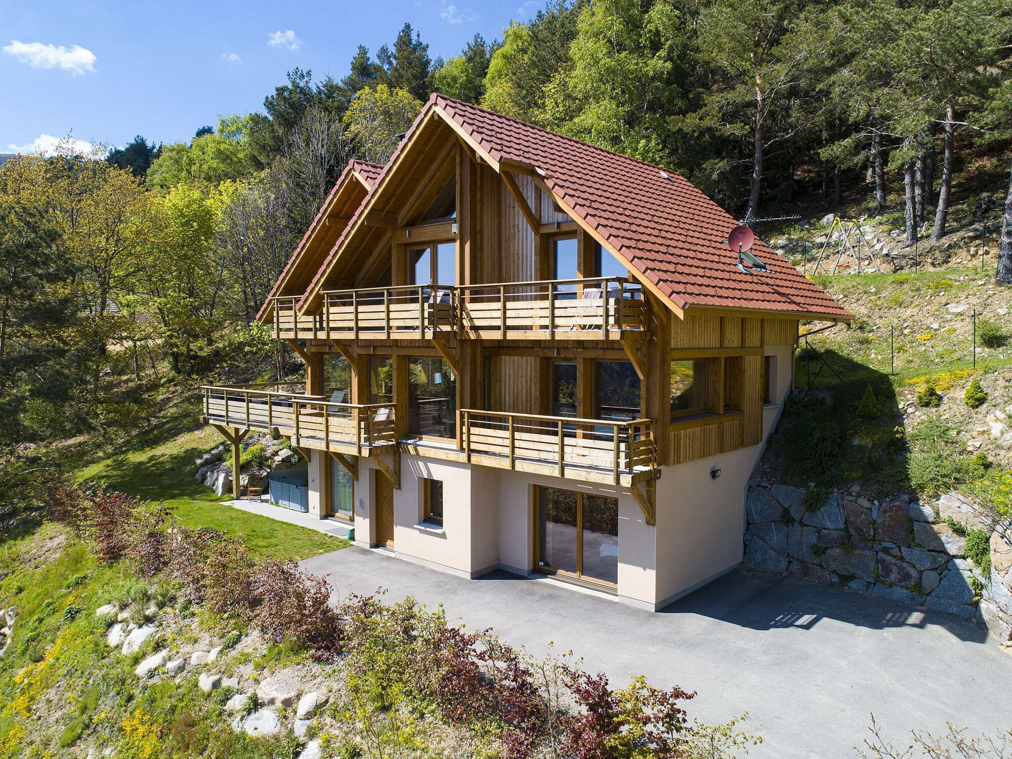 Un chalet en bois à deux étages, doté d’un balcon, situé dans une région montagneuse, entouré d’arbres et offrant une vue sur un ciel dégagé.