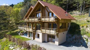 A two-story wooden chalet with a balcony, set in a mountainous area with trees and a clear sky.
