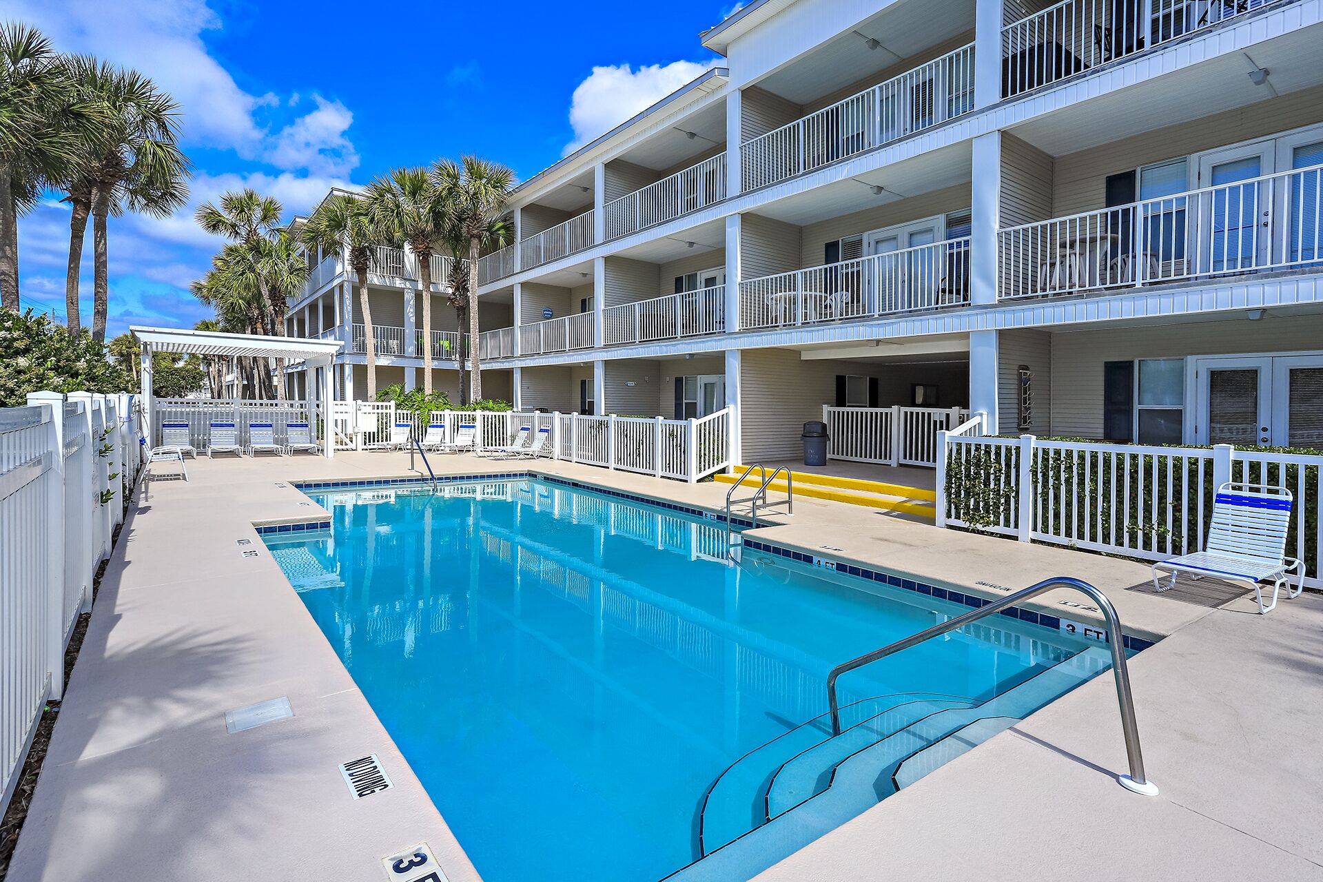 A pool area with a clear blue pool, white railings, and a multi-story building with balconies.