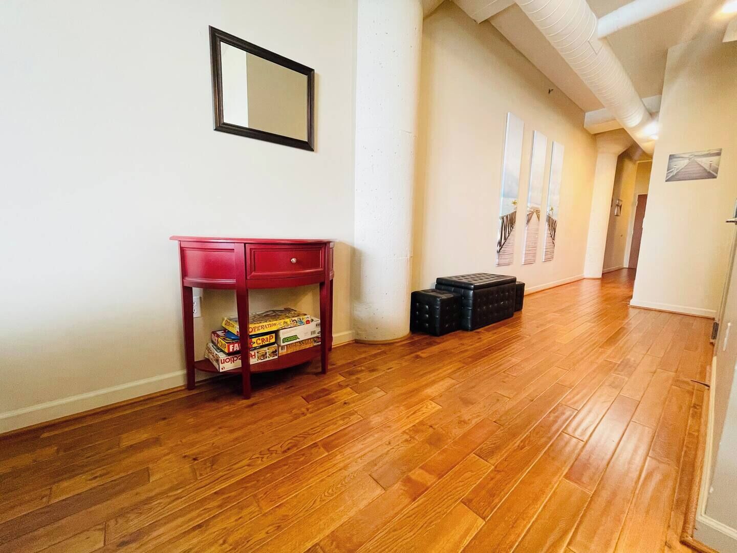 Un pasillo con suelo de madera, una consola roja con juegos de mesa, un espejo en la pared y cubos de almacenamiento negros.
