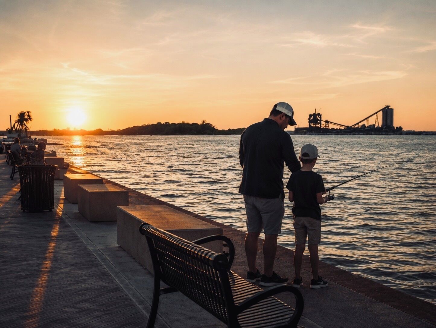 A waterfront promenade with a bench and a person fishing.