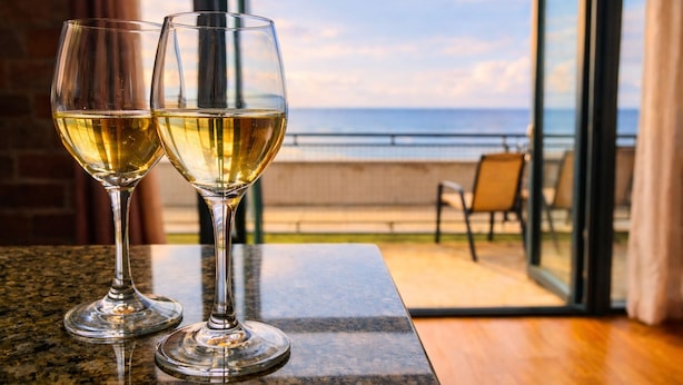 Two glasses of white wine on a table with a view of the ocean through a glass door.