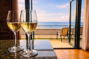 Two glasses of white wine on a table with a view of the ocean through a glass door.
