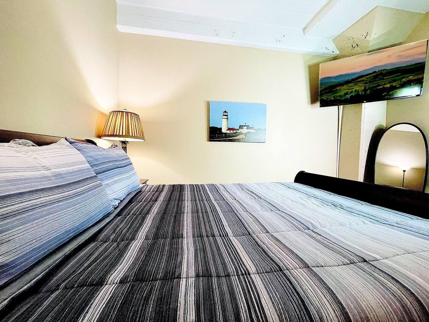 A hotel room with a bed, a lamp, a painting of a lighthouse, and a mirror.