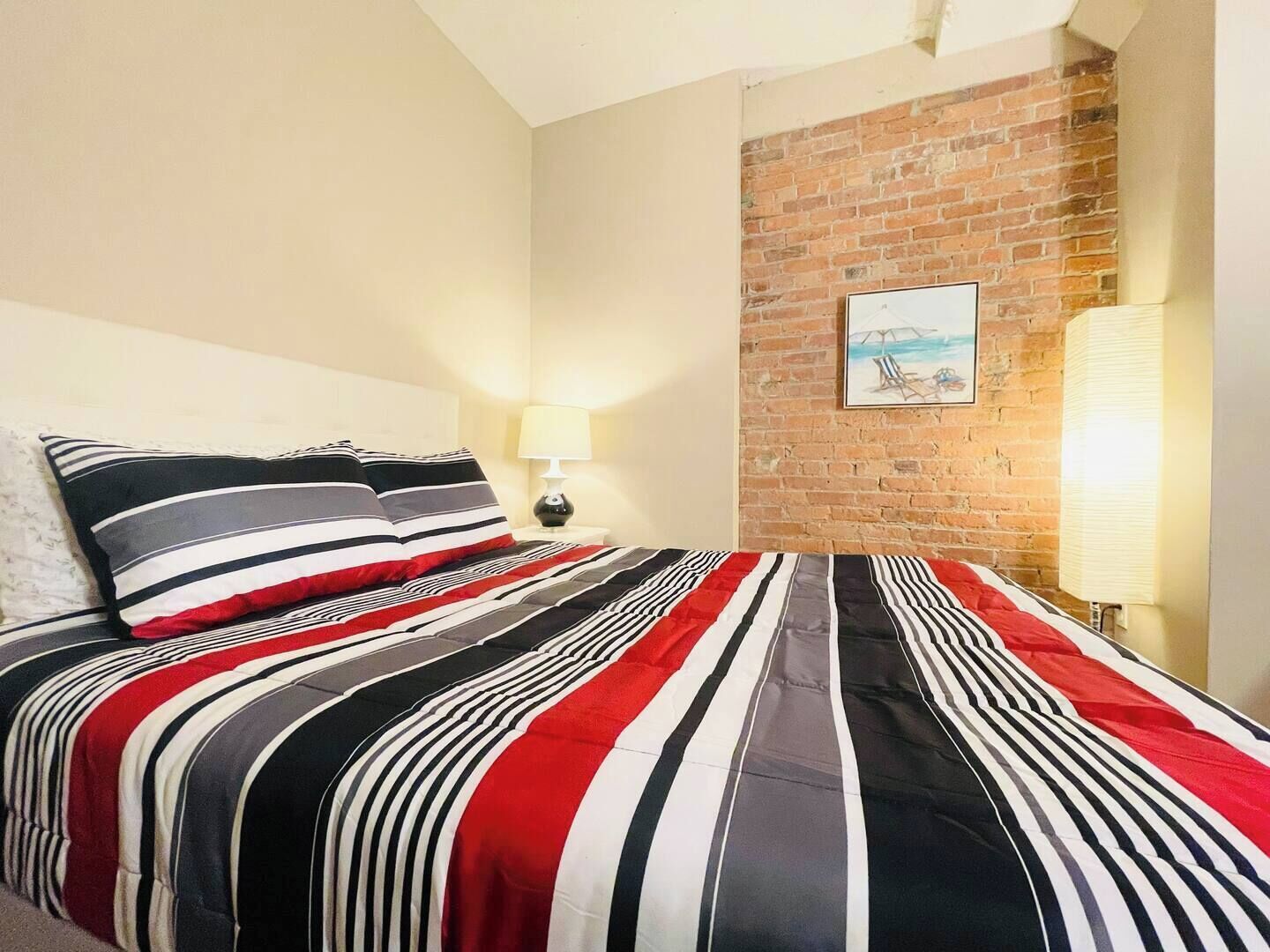 A bed with striped bedding, a lamp, and a brick wall with a picture.
