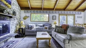 A cozy living room with a stone fireplace, a sofa, a coffee table, and a large window with a view of trees. - SS12-Bass, Blue Gill, and Catfish Pond! Hot Tub! Dolly Sods Access! (Davis)