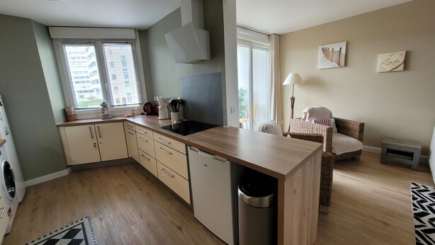 Une cuisine compacte dotée d’un lave-vaisselle intégré, un coin repas avec une chaise en osier et une vue sur l’extérieur par une fenêtre.