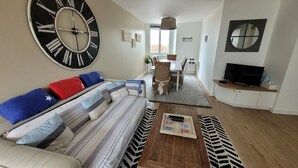 Un salon avec une grande horloge murale, un canapé orné de coussins à rayures, un coin repas avec une table et des chaises, et un téléviseur posé sur un meuble.
