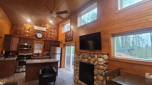 Een knus interieur van een chalet met een stenen openhaard, houten wanden, een keuken met moderne apparatuur en een grote televisie boven de openhaard.