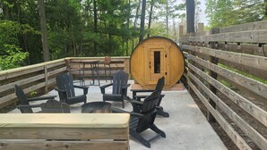 Een houten terras met Adirondack-stoelen en een grote, vatvormige constructie, omringd door een houten hek en bomen.