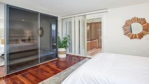 Un dormitorio con puerta corrediza de vidrio que da a un balcón, cama con ropa de cama blanca y un espejo con diseño de sol en la pared.