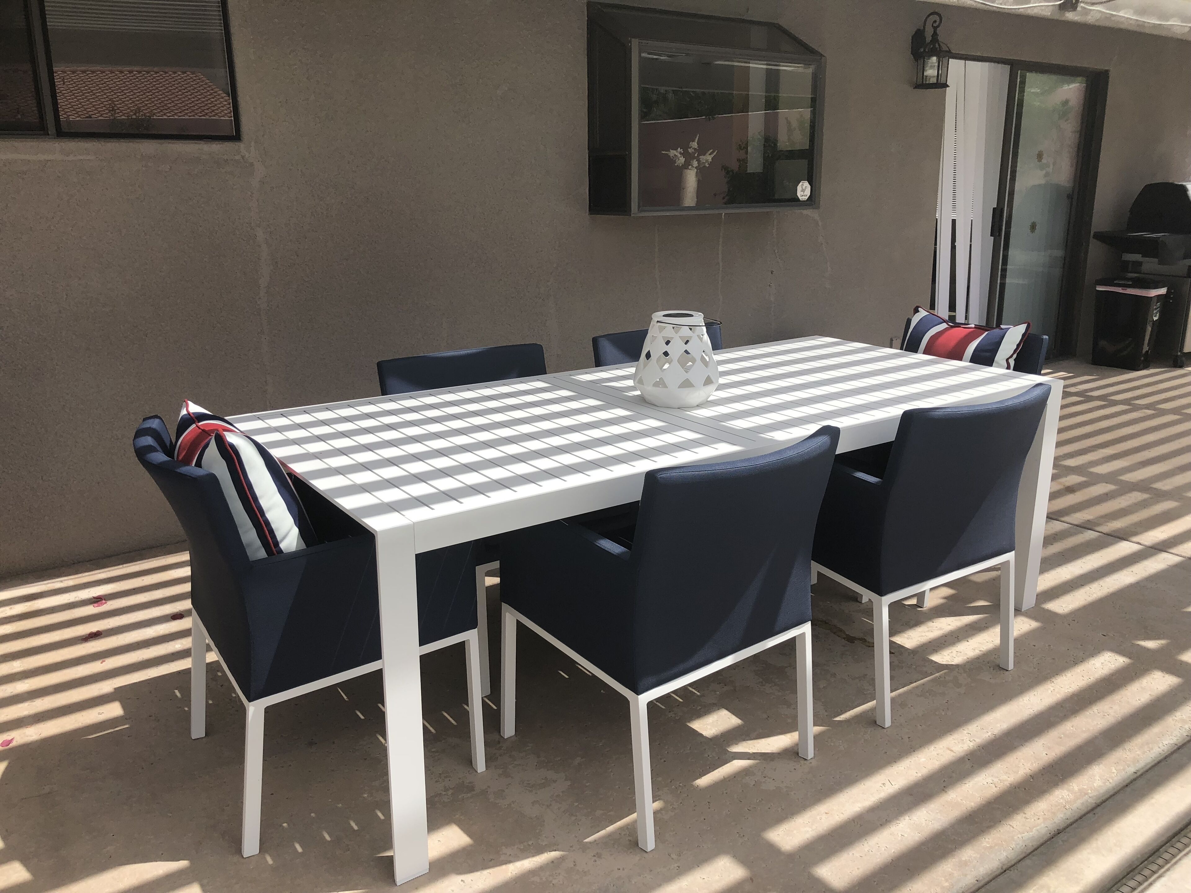 An outdoor patio with a white table and navy blue chairs, a white vase, and a black grill.