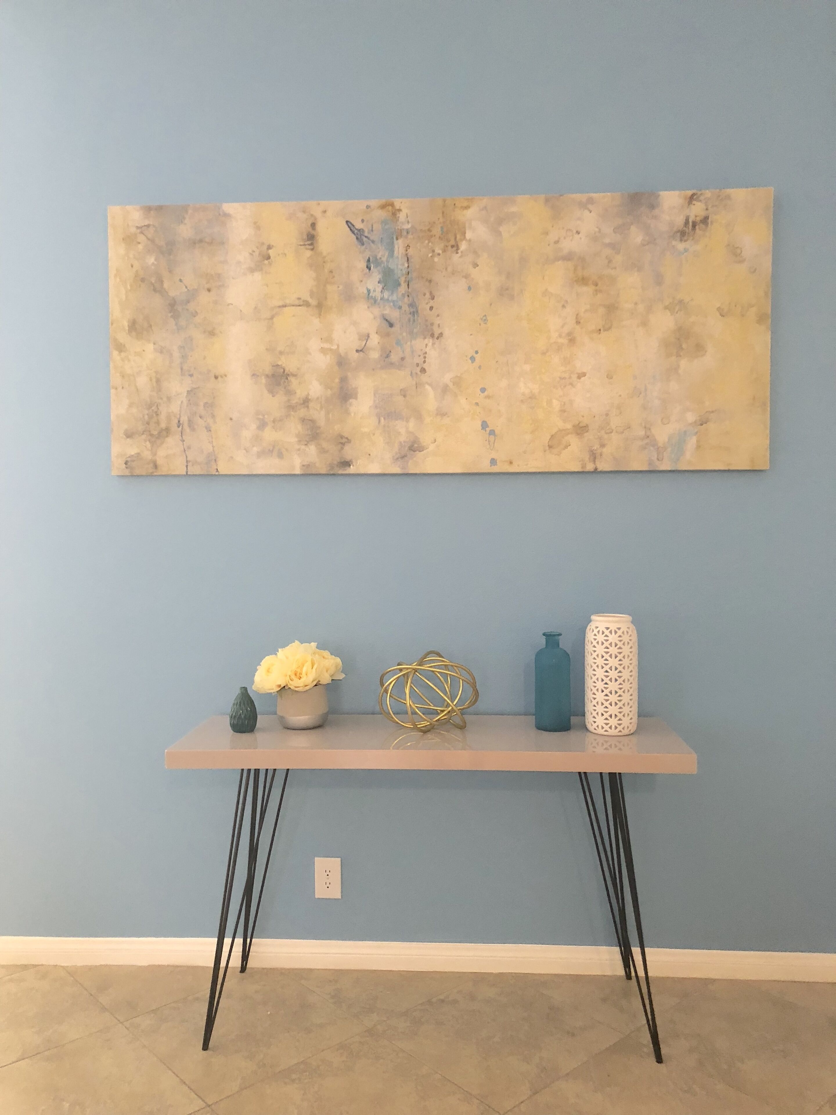 Een kamer met een blauwe wand, een lichte houten tafel met zwarte poten, en een groot abstract schilderij.