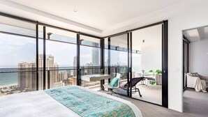 A modern hotel room with a large bed, a desk, and a view of the city and ocean. - Sealuxe Seaview Suites - Central Surfers Paradise (Surfers Paradise)