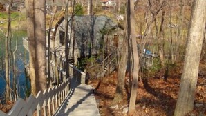 Un sendero de madera que conduce a un puente de piedra sobre un cuerpo de agua, rodeado de árboles y un edificio distante.
