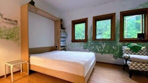 Un dormitorio con una cama de madera, una mesita de noche pequeña y una silla con un cojín estampado. Hay tres ventanas que ofrecen vistas a zonas verdes.