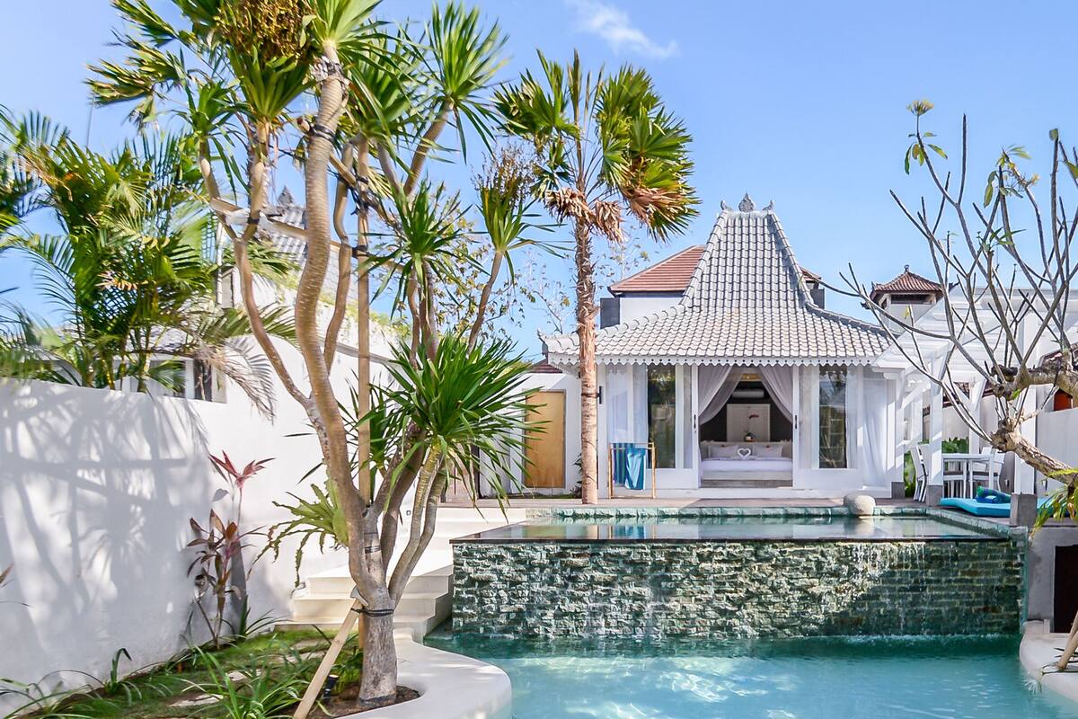 A pool with a waterfall feature, surrounded by palm trees and a traditional building with a tiled roof.