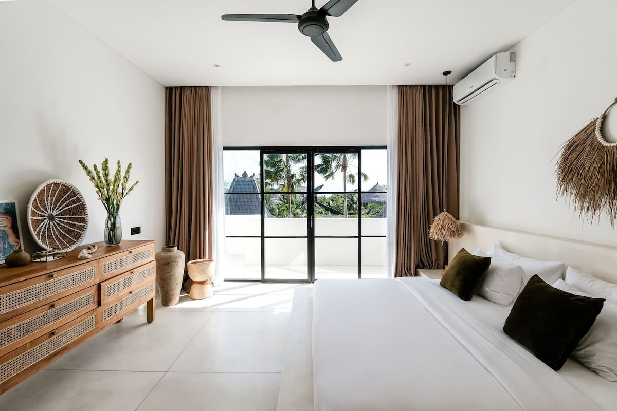 A modern hotel room with a large bed, a wooden dresser, a ceiling fan, and a view of palm trees through the window.
