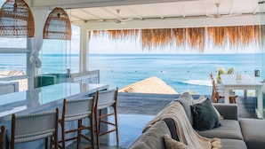 A modern room with a glass wall overlooking the ocean, featuring a sofa, chairs, and a dining table. - Mahi Mahi Villa & Suites (Pecatu)