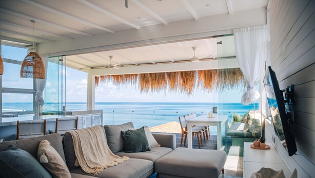 A modern living room with a large window overlooking the ocean, a sofa, a dining area, and ceiling fans. - Mahi Mahi Villa & Suites (Pecatu)