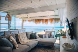 A modern living room with a large window overlooking the ocean, a sofa, a dining area, and ceiling fans. - Mahi Mahi Villa & Suites (Pecatu)