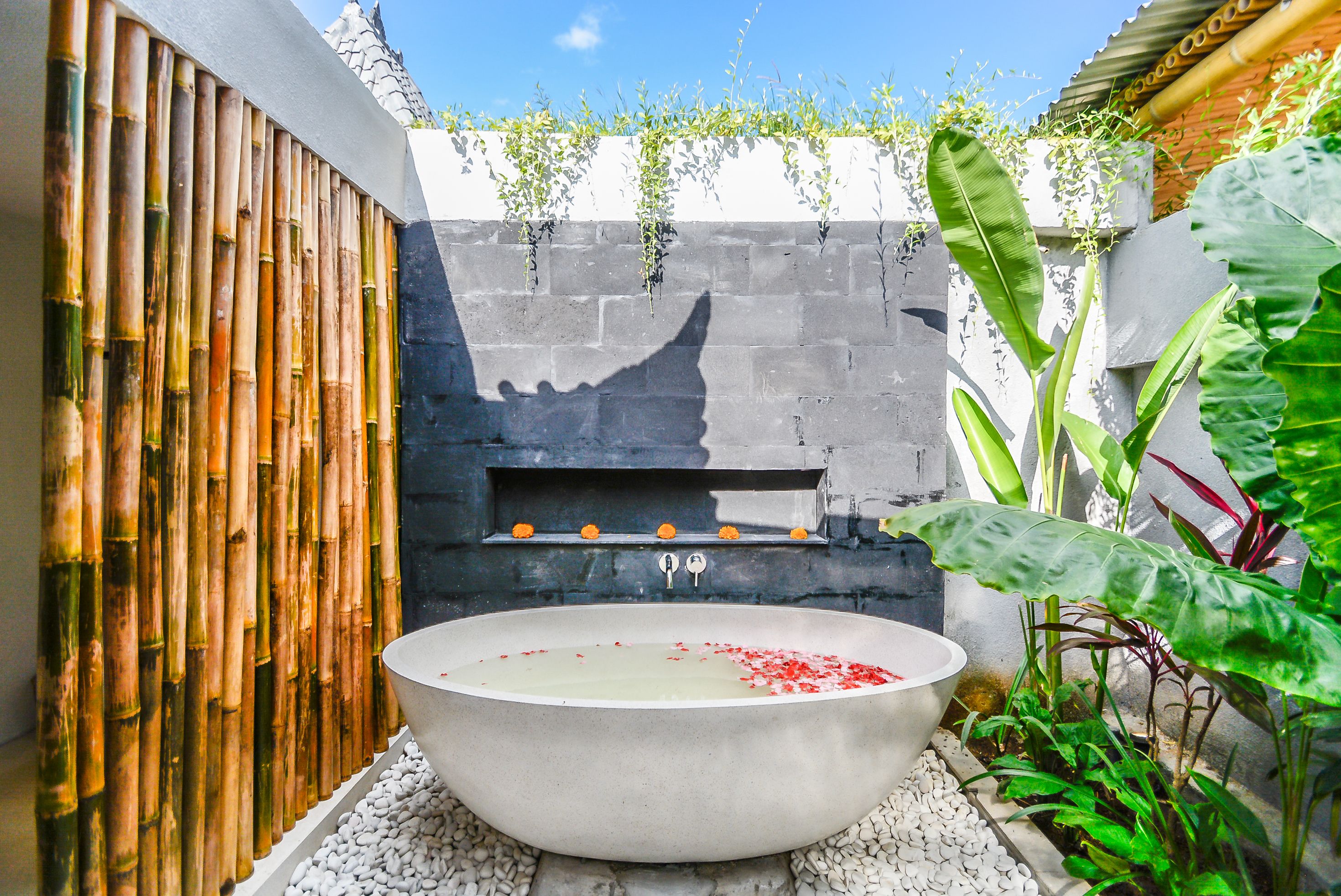 Eine Steinebadewanne mit roten Blütenblättern, umgeben von weißen Kieselsteinen, mit einer Steinwand und einem Bambuszaun im Hintergrund.