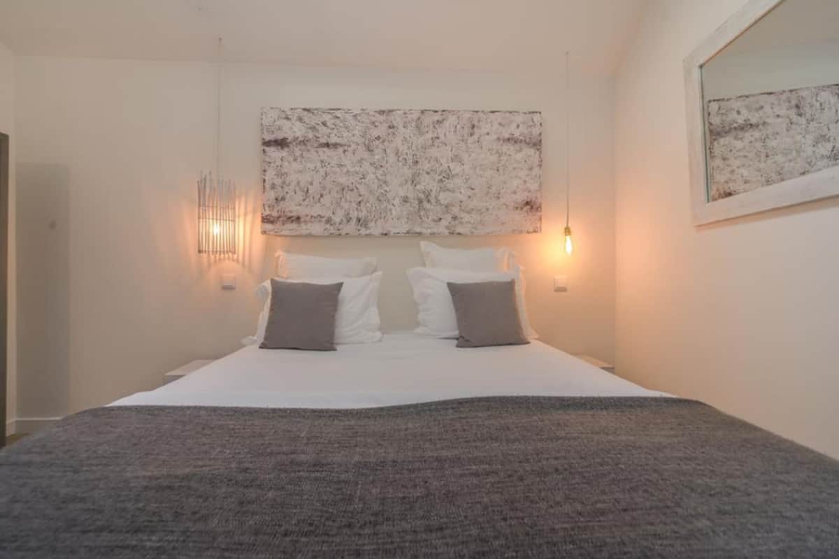 A bedroom with a large bed, two bedside lamps, and a textured artwork above the bed.