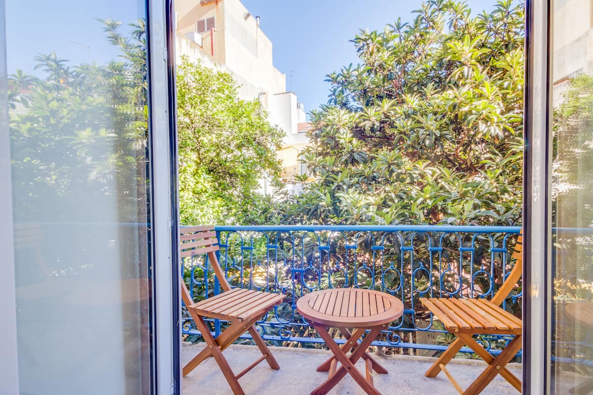 A balcony with two wooden chairs and a small round table, surrounded by a blue metal railing and lush greenery.