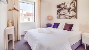 A hotel room with a large bed, a bedside table, a hanging light fixture, a painting on the wall, and a window with curtains. - Chiado 44 (Lisbon)