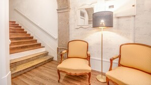 A staircase with wooden steps and white walls, two upholstered chairs with a light-colored fabric, and a floor lamp with a patterned shade. - Chiado 44 (Lisbon)