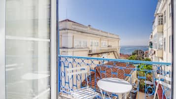 Un balcone con tavolo e sedie bianche, affacciato su una città costiera con edifici storici e vista sul mare.