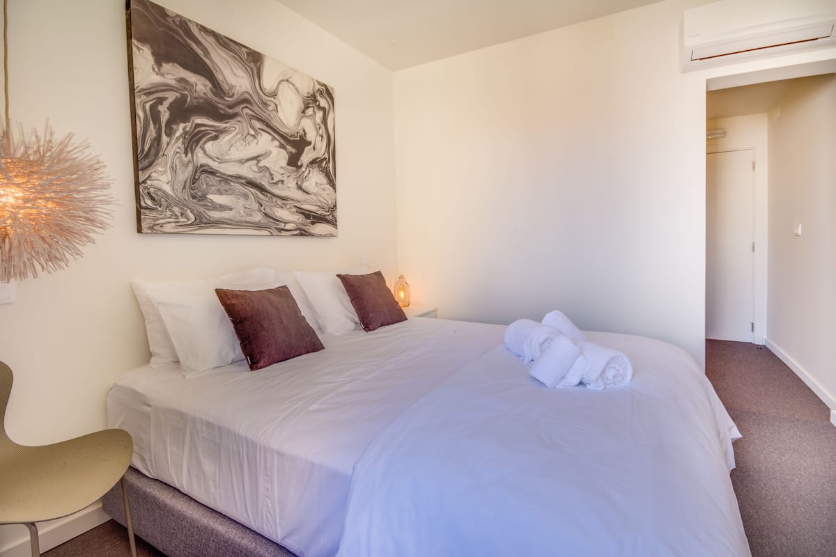 A neatly made bed with white and brown pillows, a large abstract painting on the wall, and a chair with a unique light fixture.