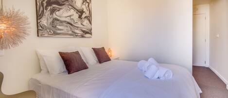 Una cama bien hecha con almohadas blancas y marrones, un cuadro abstracto grande en la pared y una silla con una lámpara de diseño original.